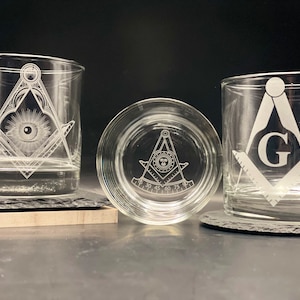 May include: Three clear, engraved drinking glasses with Masonic symbols. One glass features an eye within a triangle, another has the letter "G", and the third displays a compass and square. The glasses are on coasters.