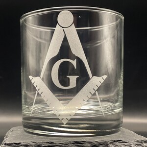 May include: Clear glass tumbler featuring a Masonic symbol, including a compass, square, and the letter "G". The design is etched into the glass. The tumbler is displayed on a dark gray surface against a black background.