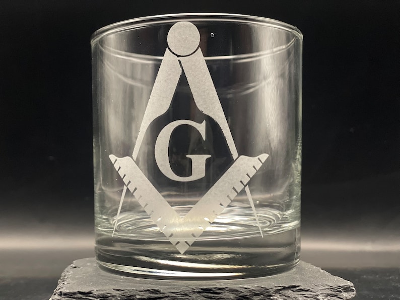 May include: Clear glass tumbler featuring a Masonic symbol, including a compass, square, and the letter "G". The design is etched into the glass. The tumbler is displayed on a dark gray surface against a black background.