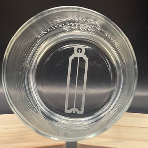 May include: Clear glass tumbler with etched design. The design includes a vertical rectangular shape with a rounded top and bottom, and the text "Brothers 2023". The glass is round and sits on a wooden surface.