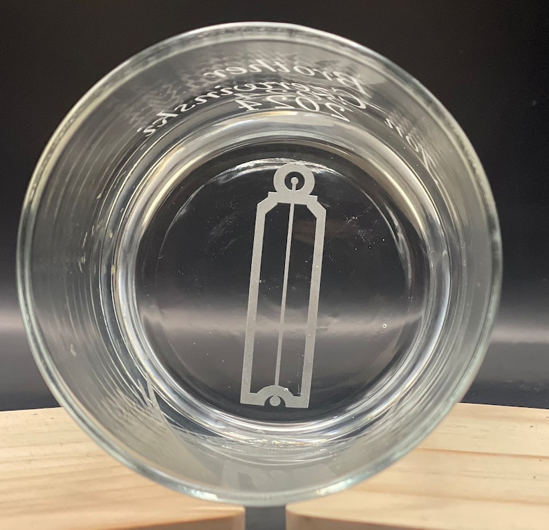 May include: Clear glass tumbler with etched design. The design includes a vertical rectangular shape with a rounded top and bottom, and the text "Brothers 2023". The glass is round and sits on a wooden surface.