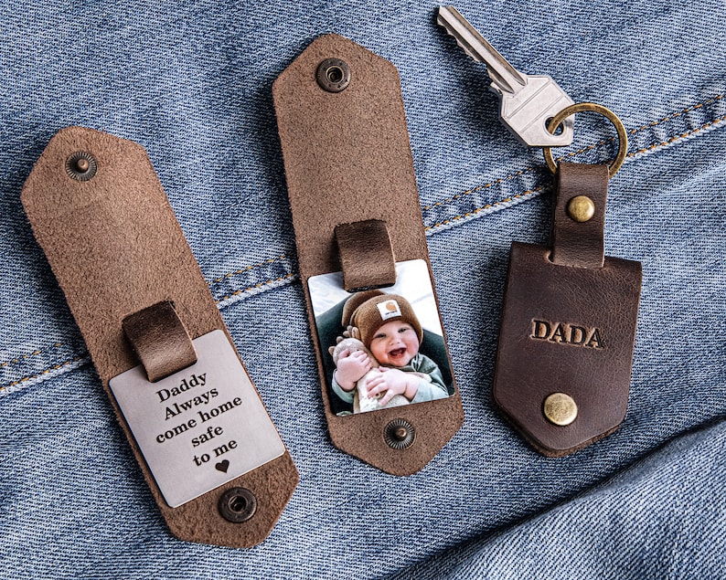 May include: Two brown leather keychains with a silver metal insert. One keychain has a photo of a baby boy and the text "Dada". The other keychain has the text "Daddy Always come home safe to me".