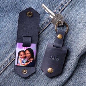 以下が含まれることがあります： 写真入れと「DAD」の刻印が施された濃い青色の革製のキーホルダー。写真入れには、母親と娘の姿が写っています。