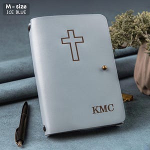 Puede incluir: Diario de cuero azul hielo con una cruz y las iniciales "KMC". El diario tiene un cierre de bot&oacute;n dorado. Hay un bol&iacute;grafo negro al lado. El texto "M-size ICE BLUE" est&aacute; en la esquina superior izquierda.