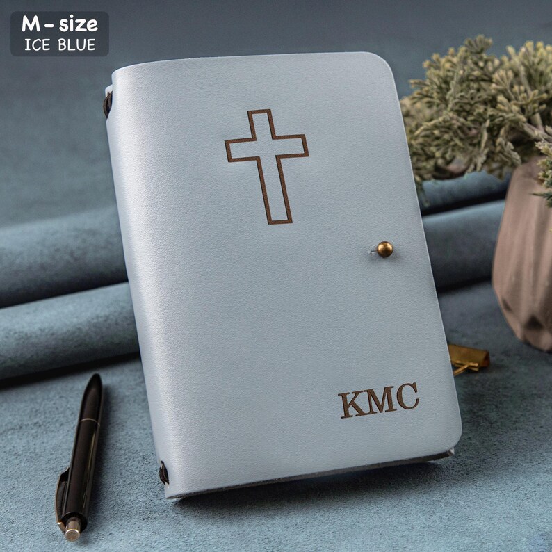 Puede incluir: Diario de cuero azul hielo con una cruz y las iniciales "KMC". El diario tiene un cierre de bot&oacute;n dorado. Hay un bol&iacute;grafo negro al lado. El texto "M-size ICE BLUE" est&aacute; en la esquina superior izquierda.