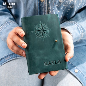 Puede incluir: Un diario de cuero verde azulado con un dise&ntilde;o de rosa de los vientos en relieve en la portada. El nombre "KAYLA" est&aacute; grabado debajo. El diario se cierra con un peque&ntilde;o bot&oacute;n de color lat&oacute;n.