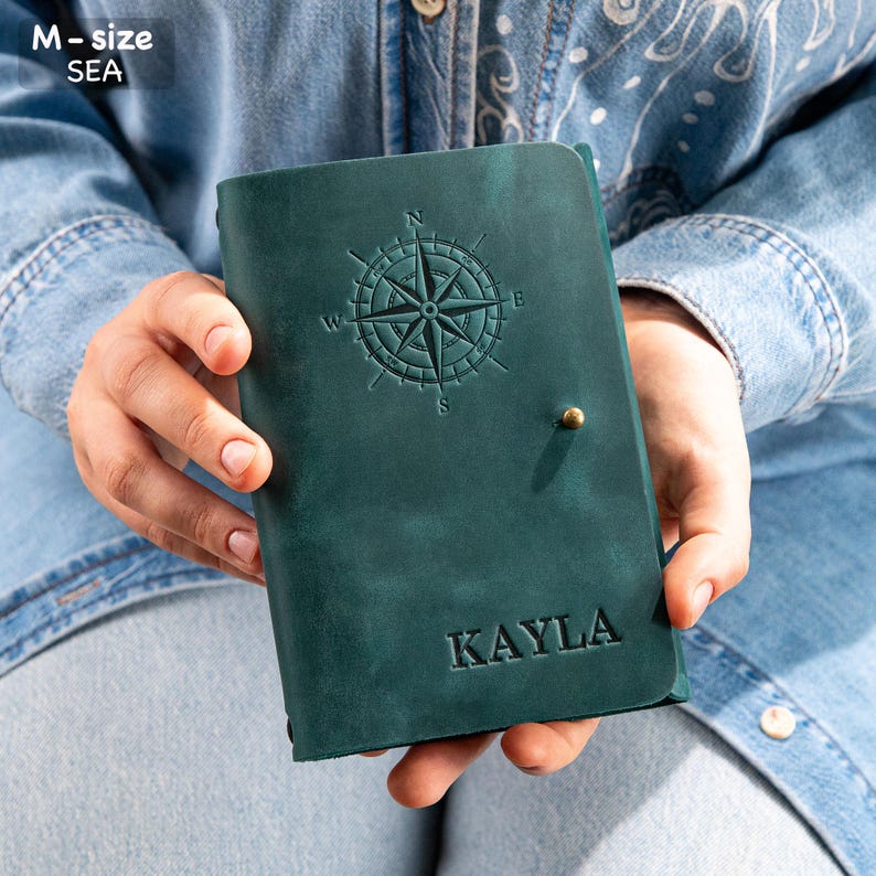 Puede incluir: Un diario de cuero verde azulado con un dise&ntilde;o de rosa de los vientos en relieve en la portada. El nombre "KAYLA" est&aacute; grabado debajo. El diario se cierra con un peque&ntilde;o bot&oacute;n de color lat&oacute;n.