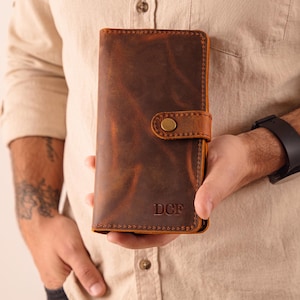 May include: A rectangular brown leather wallet featuring a distressed finish, visible stitching along the edges, and a snap-button closure tab. The lower right corner is embossed with the capital letters DCF in a serif font.