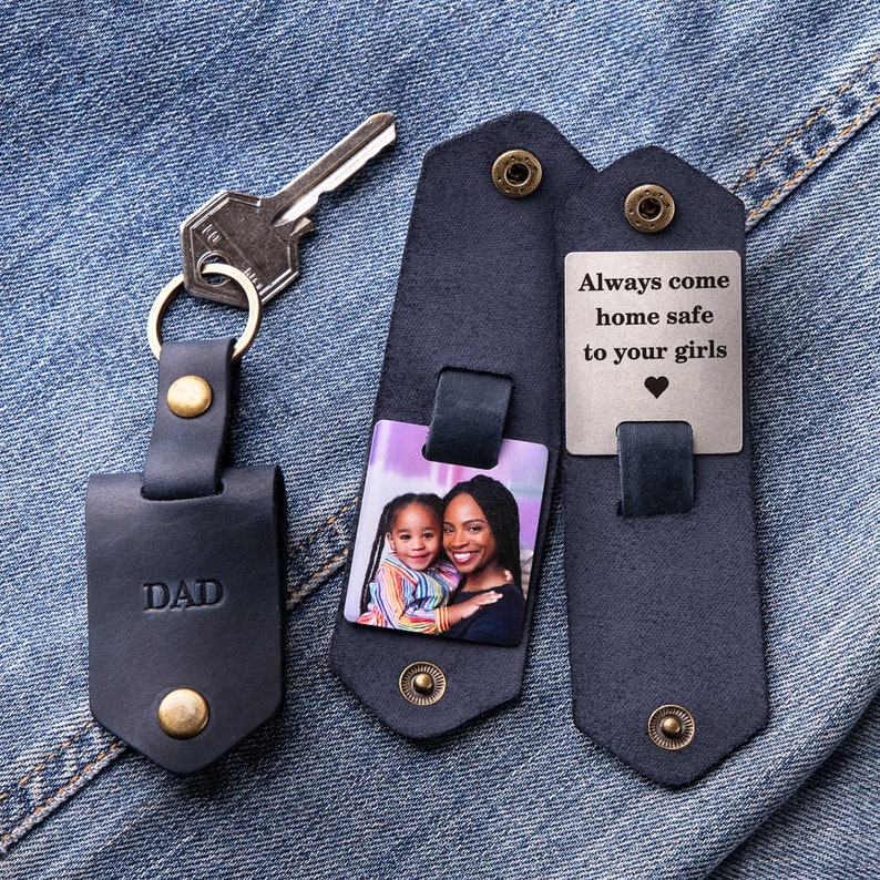 May include: A black leather keychain with a photo insert and a metal tag that reads "Always come home safe to your girls". The photo insert shows a young girl and a woman smiling. The keychain also has a metal tag that reads "DAD".