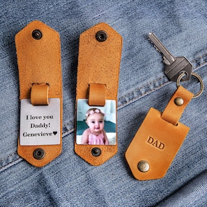 May include: Two brown leather keychains with a metal insert. One keychain has a photo of a baby girl wearing a pink dress and a flower crown. The other keychain has the text "I love you Daddy! Genevieve" and a heart. The third keychain is brown leather with the word "DAD" embossed on it.