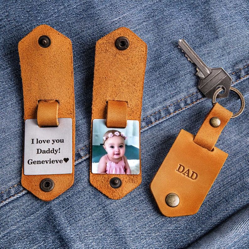 May include: Two brown leather keychains with a metal insert. One keychain has a photo of a baby girl wearing a pink dress and a flower crown. The other keychain has the text "I love you Daddy! Genevieve" and a heart. The third keychain is brown leather with the word "DAD" embossed on it.
