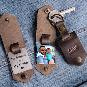 May include: Two brown leather keychains with metal inserts. One keychain has a photo of a man and a child, and the other has the text "My biggest hero My Daddy" with a heart symbol. The keychain with the photo also has the initials "SBW" engraved on the back.
