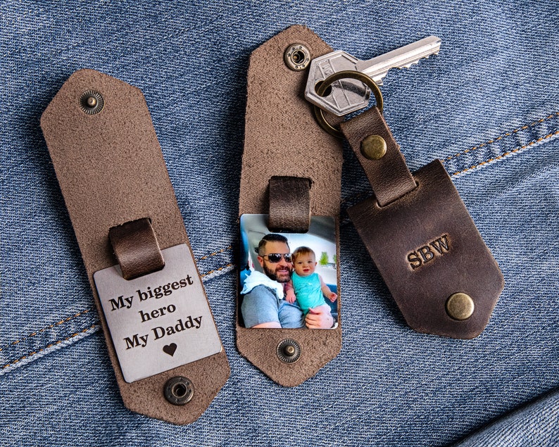May include: Two brown leather keychains with metal inserts. One keychain has a photo of a man and a child, and the other has the text "My biggest hero My Daddy" with a heart symbol. The keychain with the photo also has the initials "SBW" engraved on the back.