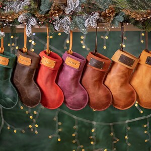 May include: A row of personalized leather Christmas stockings in various colors, including green, brown, red, and purple. Each stocking has a leather tag with a name engraved on it. The stockings are hanging from a wooden shelf decorated with greenery and lights.