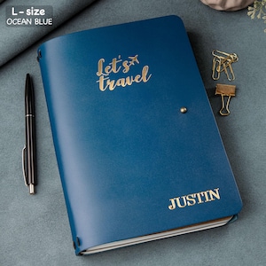May include: Ocean blue L-size journal with the words "Let's Travel" and a small airplane graphic in gold lettering. The name "JUSTIN" is also in gold lettering. A black pen and gold paper clips are next to the journal.