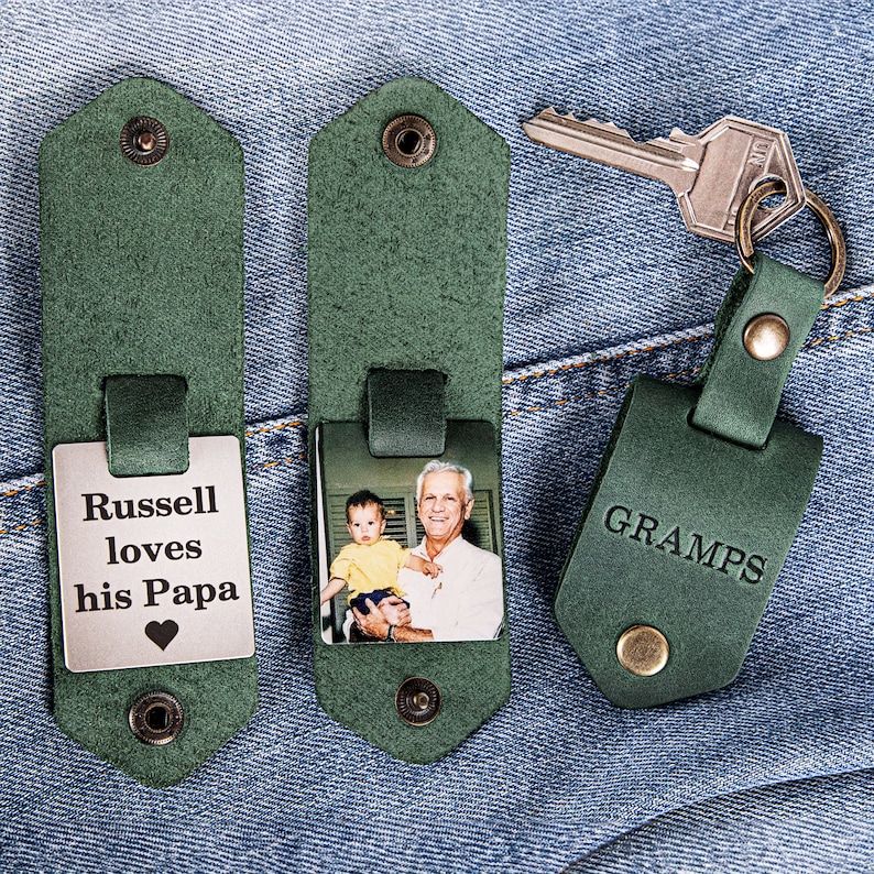 May include: Two green leather keychains with a photo insert and a metal plate. One keychain has a photo of a man and a child, and the other has a metal plate with the text "Russell loves his Papa" and a heart. The third keychain is green leather with the word "GRAMPS" embossed on it.