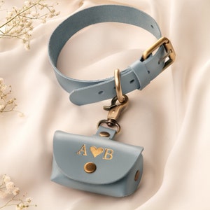May include: A light blue leather dog collar with a matching waste bag holder. The collar has a brass buckle and a D-ring. The waste bag holder is embossed with the letters "A" and "B" with a heart symbol in gold. The holder has a snap closure.