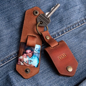 May include: A brown leather keychain with a photo insert and a separate leather tag that says "DAD". The photo insert shows a man holding a young child.