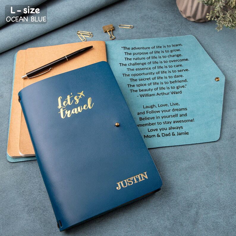 Puede incluir: Diario azul oc&eacute;ano tama&ntilde;o L con el texto "Let's travel" en dorado. El diario est&aacute; personalizado con el nombre "JUSTIN". Se ve un bol&iacute;grafo y papel. El diario est&aacute; abierto, revelando texto inspirador. Tema: viajes.