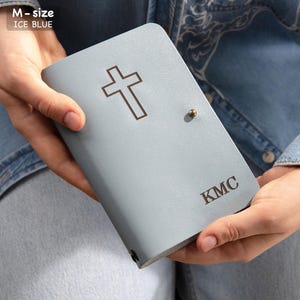 Może przedstawiać: Błękitna okładka Biblii z krzyżem i inicjałami "KMC". Okładka ma zapięcie na guzik i tekst "M-size ICE BLUE" w lewym g&oacute;rnym rogu. Okładka jest trzymana w dłoniach.