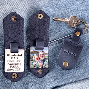 May include: Two black leather keychains with a photo insert. One keychain has a photo of a man and a baby. The other keychain has the text "Wonderful Dad since 1991 Awesome Papa since 2021". The third keychain is black leather with the word "POPPY" in white text.