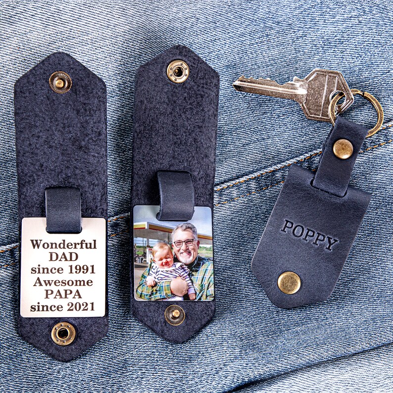 May include: Two black leather keychains with a photo insert. One keychain has a photo of a man and a baby. The other keychain has the text "Wonderful Dad since 1991 Awesome Papa since 2021". The third keychain is black leather with the word "POPPY" in white text.