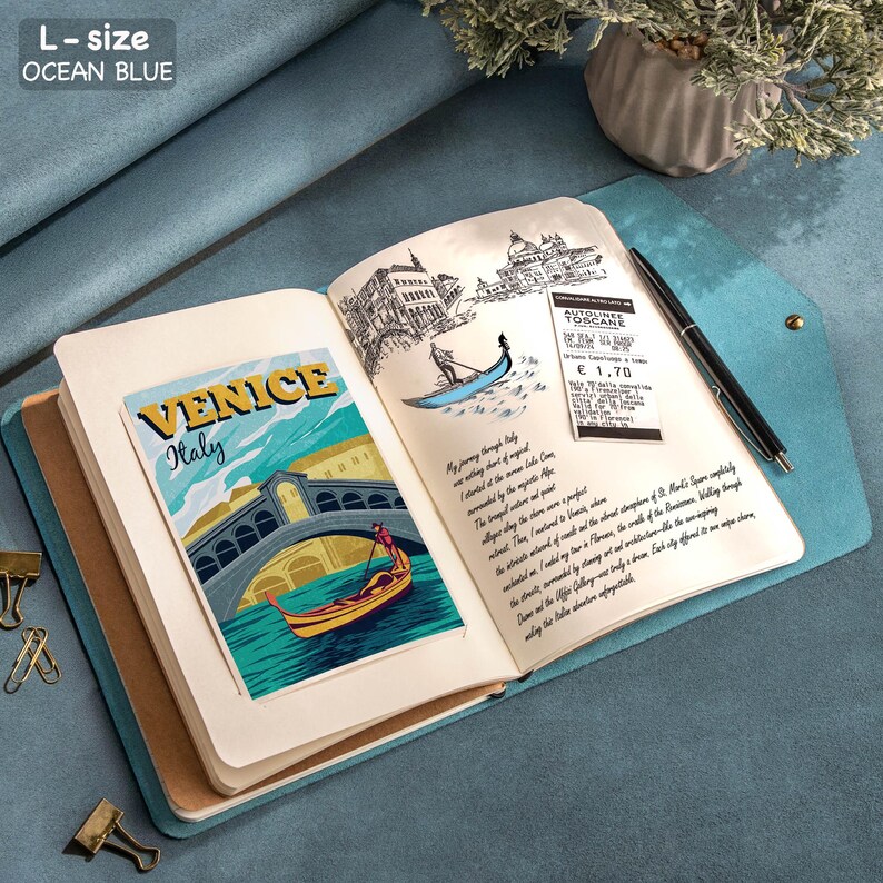Puede incluir: Un diario azul oc&eacute;ano abierto con el texto "L-size OCEAN BLUE" en la parte superior. El diario presenta una colorida postal de Venecia, Italia y bocetos de Venecia. Un bol&iacute;grafo negro descansa en la p&aacute;gina derecha.
