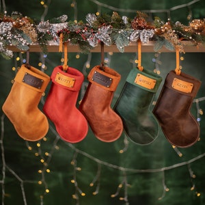 May include: Five leather Christmas stockings in shades of gold, red, brown, green, and dark brown hang from a wooden beam. Each stocking has a leather name tag with names like "Avery," "Kitty," and "Tommy." The background features festive greenery and lights.