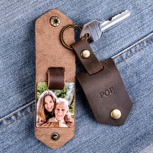 May include: A brown leather keychain with a photo insert featuring a woman and a man. The keychain has a metal ring and a brown leather tag with the word "POP" embossed on it.