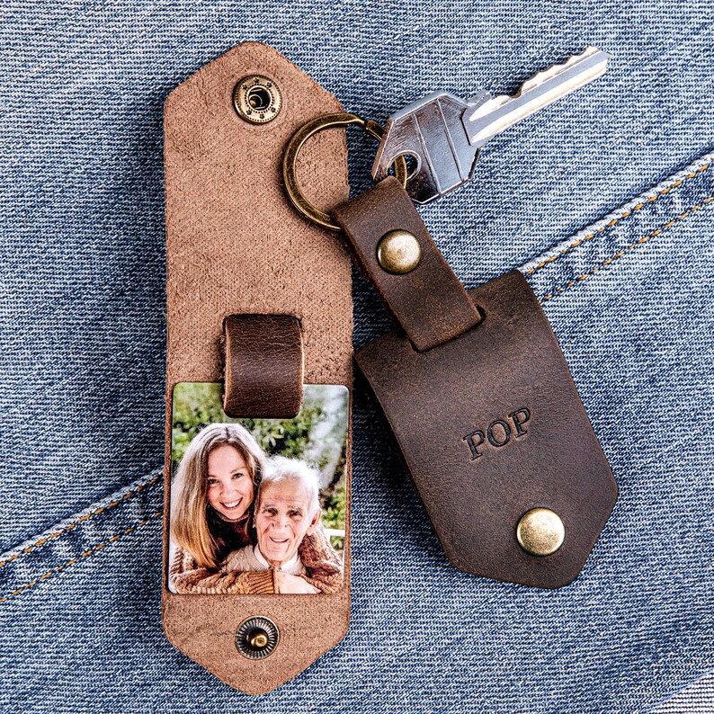 May include: A brown leather keychain with a photo insert featuring a woman and a man. The keychain has a metal ring and a brown leather tag with the word "POP" embossed on it.