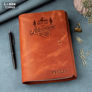 May include: A copper-colored leather journal with the text "AND SO THE ADVENTURE BEGINS" and the name "PHILIS" engraved on the cover. A black pen rests beside the closed journal. The journal is L-size.