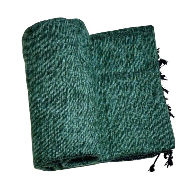 Op de afbeelding: Een opgerolde, getextureerde, groene deken met een franje van zwarte kwastjes. De deken is gemaakt van een geweven materiaal en de kleur is een diepe, rijke tint groen. De deken is over zichzelf gevouwen.