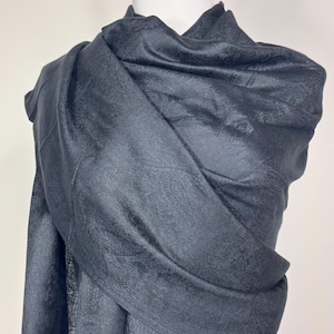 May include: A dark gray scarf draped over a mannequin. The scarf has a subtle, textured pattern and is wrapped around the mannequin's neck and shoulders. The fabric appears soft and flowing, with a slight sheen.