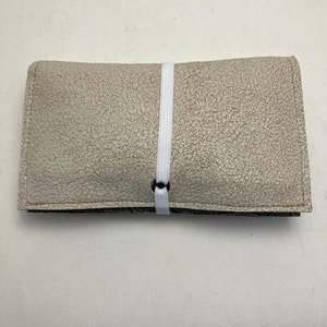 Könnte beinhalten: Eine hellgraue Ledertasche mit einem weißen Gummibandverschluss. Die Tasche hat eine strukturierte Oberfläche und ein dezentes Muster.