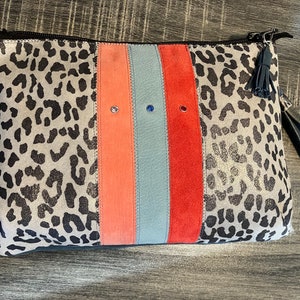 Könnte beinhalten: Eine graue und schwarze Clutch mit Leopardenmuster, Reißverschluss und schwarzem Quastenanhänger. Die Clutch hat drei vertikale Streifen in Koralle, Hellblau und Rot.
