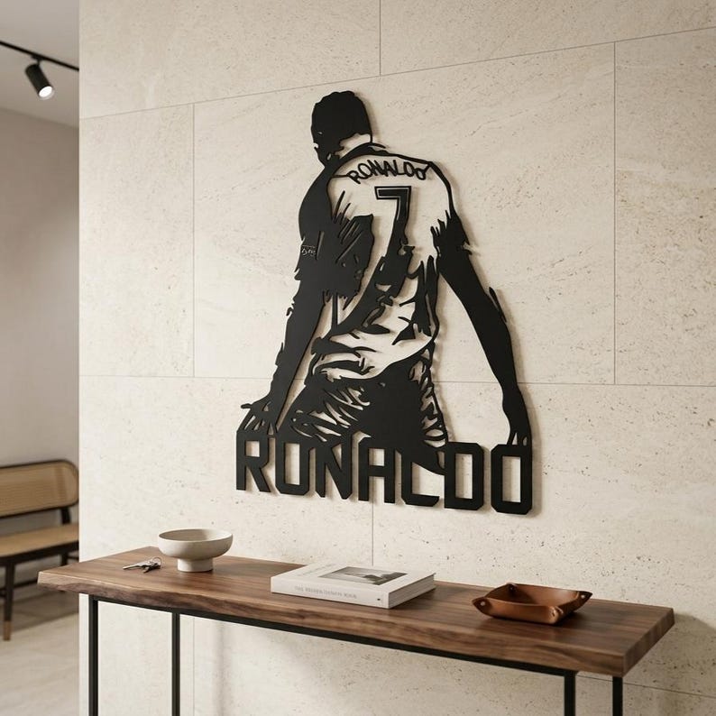 Op de afbeelding: Zwart metalen wandkunstwerk met een voetballer in een shirt met de naam "RONALDO" en het nummer "7". Het kunstwerk is bevestigd aan een lichtgekleurde muur boven een houten console.