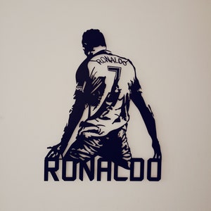 Op de afbeelding: Een zwart-wit wandkunstwerk met een silhouet van een voetballer in een shirt met de naam "RONALDO" en het nummer "7". De speler staat in een dynamische pose, met uitgestrekte armen. Het kunstwerk is op een witte muur gemonteerd.