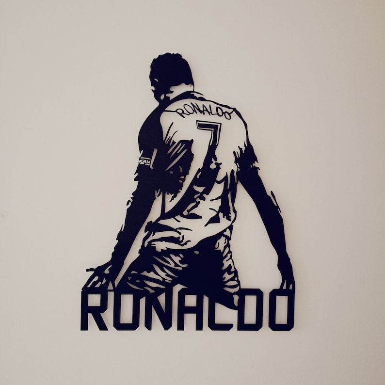 Op de afbeelding: Een zwart-wit wandkunstwerk met een silhouet van een voetballer in een shirt met de naam "RONALDO" en het nummer "7". De speler staat in een dynamische pose, met uitgestrekte armen. Het kunstwerk is op een witte muur gemonteerd.