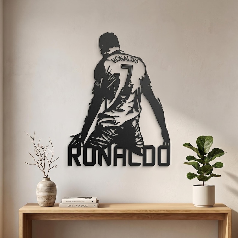 Op de afbeelding: Zwarte metalen wandkunst met een voetballer met de naam "RONALDO" eronder. De speler, van achteren gezien, draagt nummer 7. Het kunstwerk is bevestigd aan een witte muur boven een houten tafel.