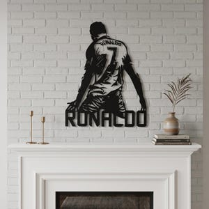 Op de afbeelding: Zwart metalen wandkunstwerk dat een knielende voetballer afbeeldt, met de naam "RONALDO" eronder. De speler draagt een shirt met het nummer "7". Het kunstwerk is bevestigd aan een witte bakstenen muur.