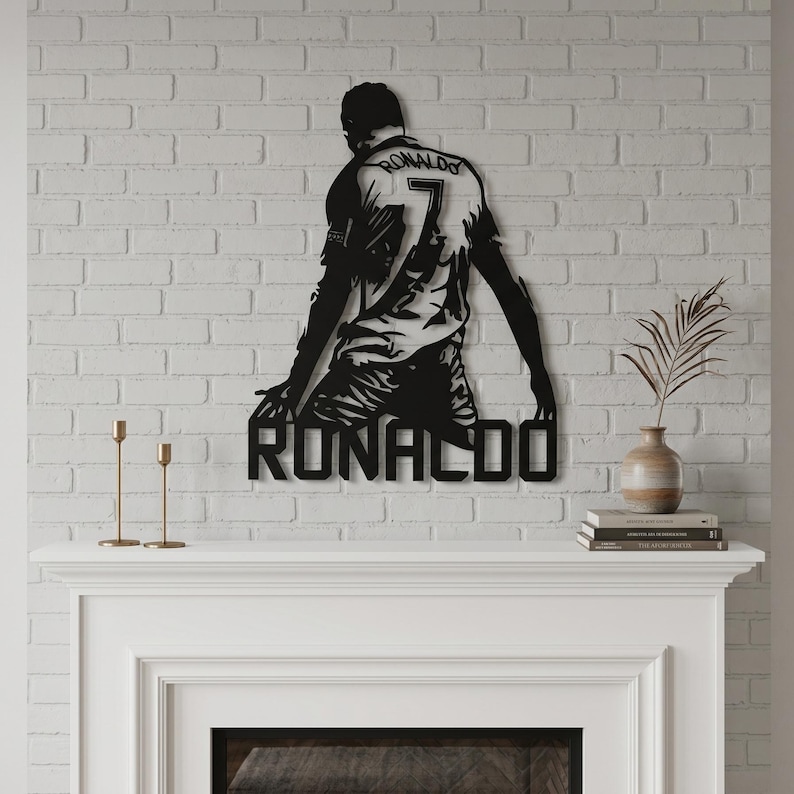Op de afbeelding: Zwart metalen wandkunstwerk dat een knielende voetballer afbeeldt, met de naam "RONALDO" eronder. De speler draagt een shirt met het nummer "7". Het kunstwerk is bevestigd aan een witte bakstenen muur.