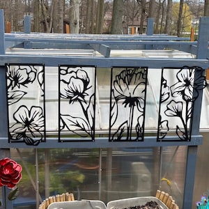 May include: Black metal floral panels with a cut-out design, mounted on a gray wooden structure. The panels feature stylized flower and leaf silhouettes. The background shows a greenhouse with a clear roof and trees.