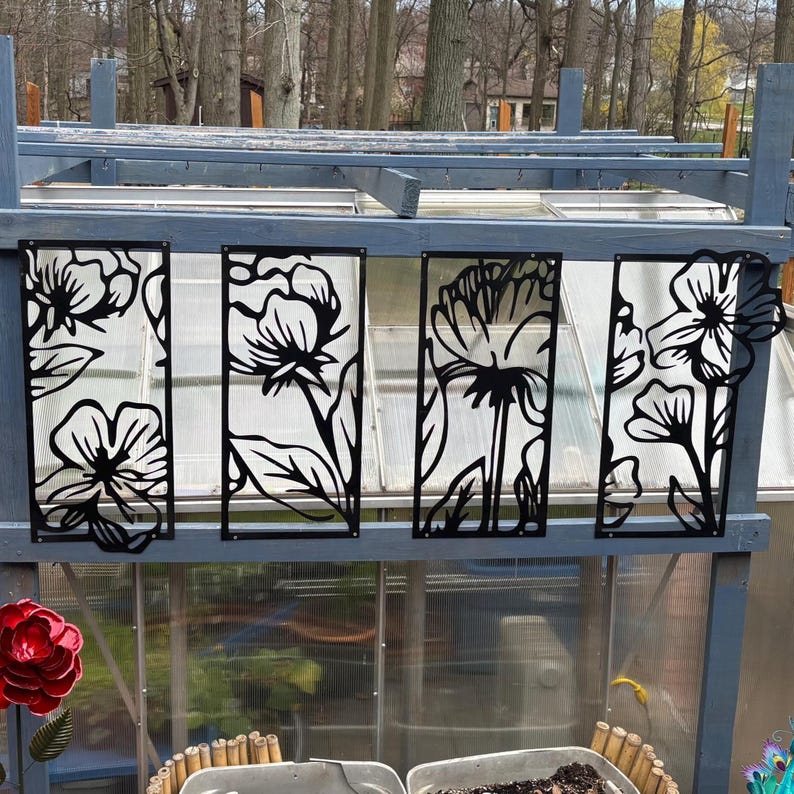 May include: Black metal floral panels with a cut-out design, mounted on a gray wooden structure. The panels feature stylized flower and leaf silhouettes. The background shows a greenhouse with a clear roof and trees.