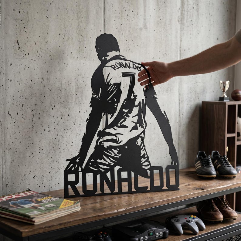 Op de afbeelding: Zwart metalen wandkunstwerk met een voetballer in een shirt met de naam "RONALDO" aan de basis en nummer 7. Het kunstwerk wordt getoond op een houten plank tegen een betonnen muur.