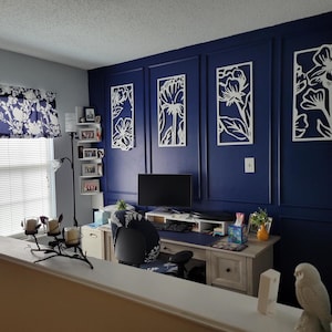 May include: A home office with a navy blue accent wall featuring white floral wall art. A desk with a computer, chair, and decor sits in front of the wall. A window with a floral valance is on the left.