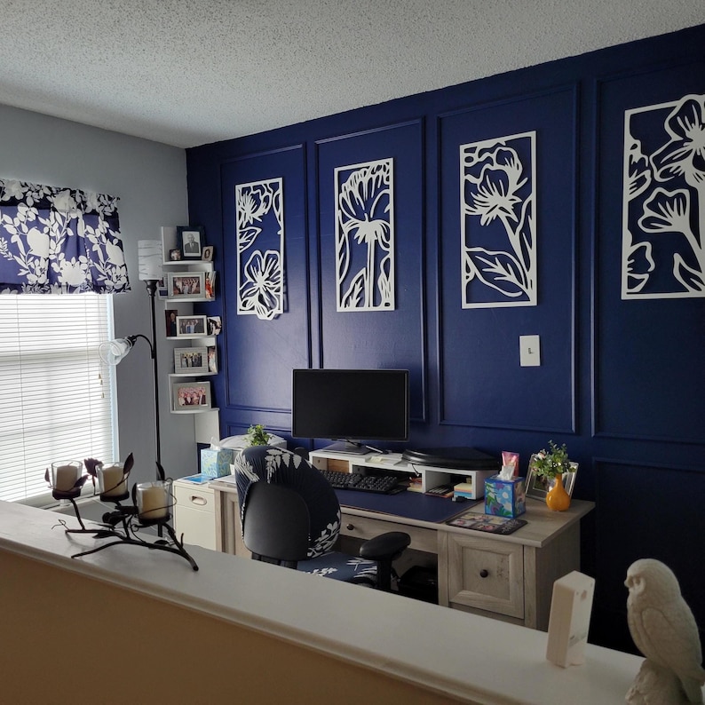 May include: A home office with a navy blue accent wall featuring white floral wall art. A desk with a computer, chair, and decor sits in front of the wall. A window with a floral valance is on the left.