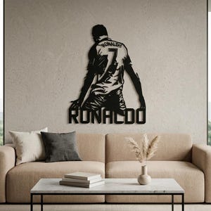 Op de afbeelding: Zwart metalen wandkunstwerk met een voetballer met nummer 7 en de naam "RONALDO". Het kunstwerk is bevestigd aan een neutrale muur boven een beige bank en salontafel.