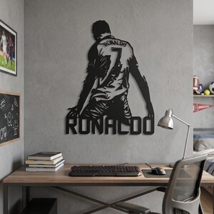Op de afbeelding: Zwart metalen wandkunst van een voetballer in een shirt met nummer 7 en de naam "RONALDO" eronder. Het kunstwerk is bevestigd aan een grijze muur, boven een houten bureau met een toetsenbord en muis.