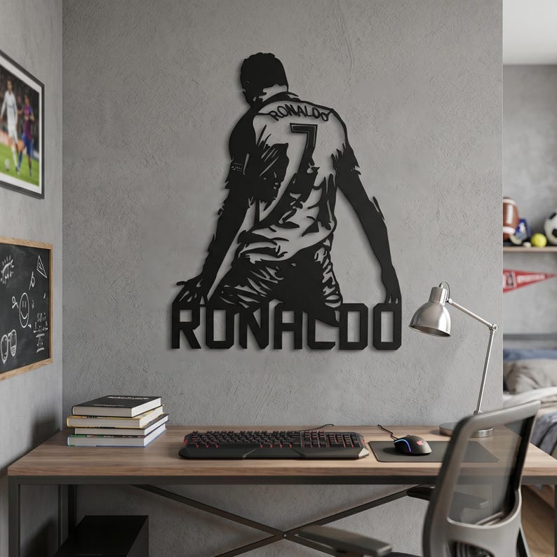 Op de afbeelding: Zwart metalen wandkunst van een voetballer in een shirt met nummer 7 en de naam "RONALDO" eronder. Het kunstwerk is bevestigd aan een grijze muur, boven een houten bureau met een toetsenbord en muis.