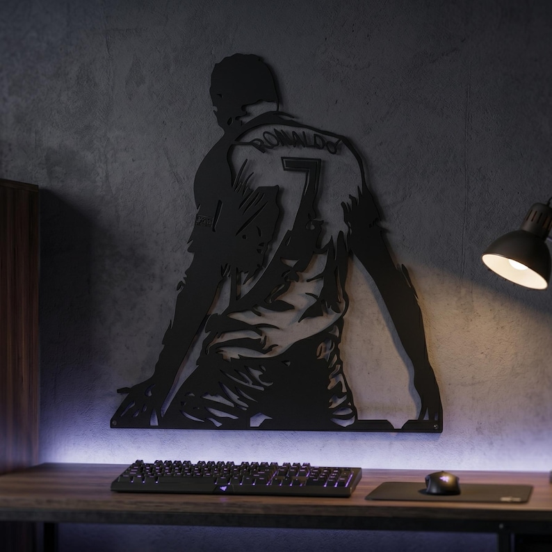 Op de afbeelding: Zwart metalen wandkunstwerk dat een voetballer in knielende pose afbeeldt, met een shirt met nummer 7 en de naam "RONALDO". Het kunstwerk is bevestigd aan een grijze muur boven een bureau met een toetsenbord en muis.